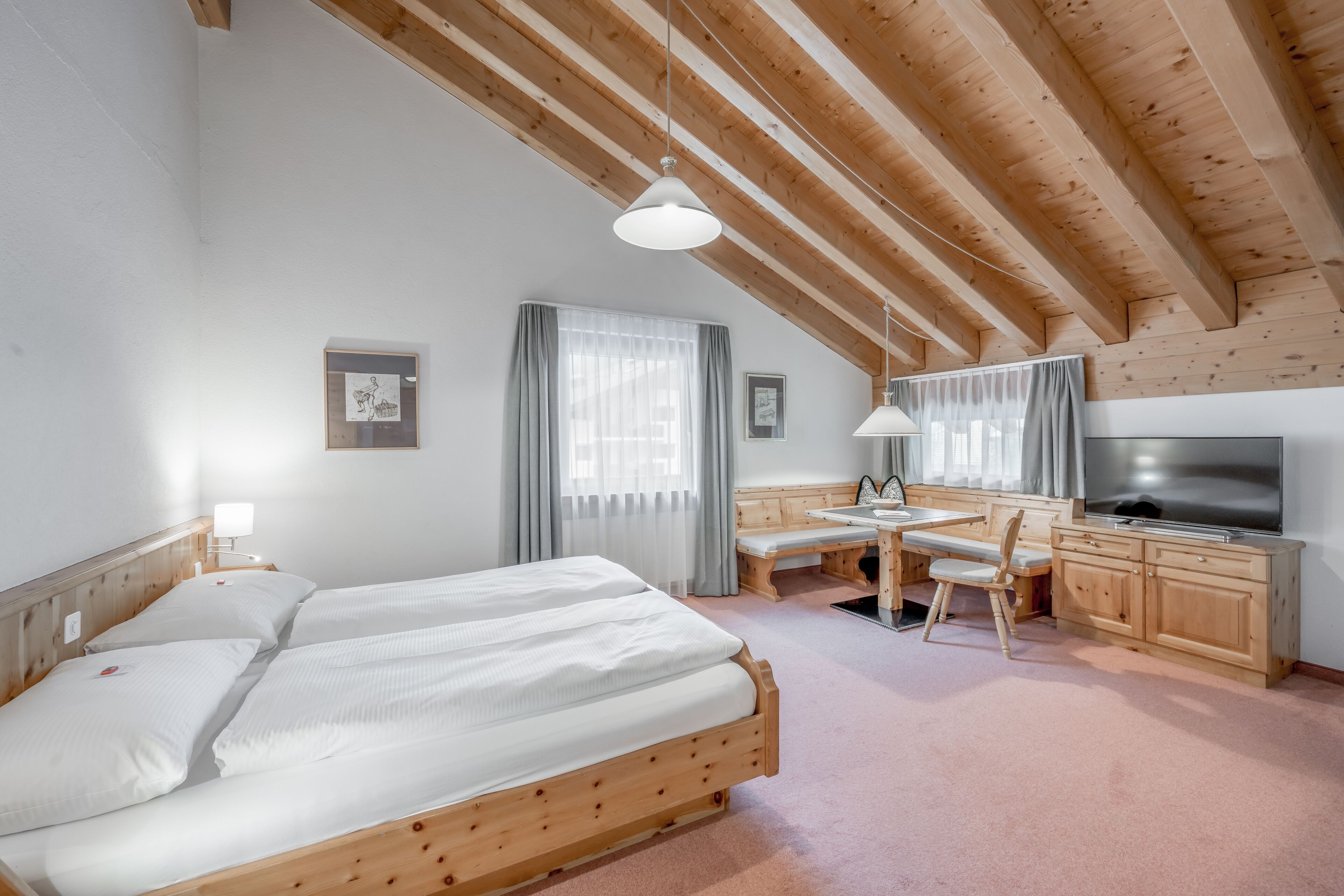 A room with a wooden ceiling, a bed, a desk, a chair, a TV, and a window with curtains.