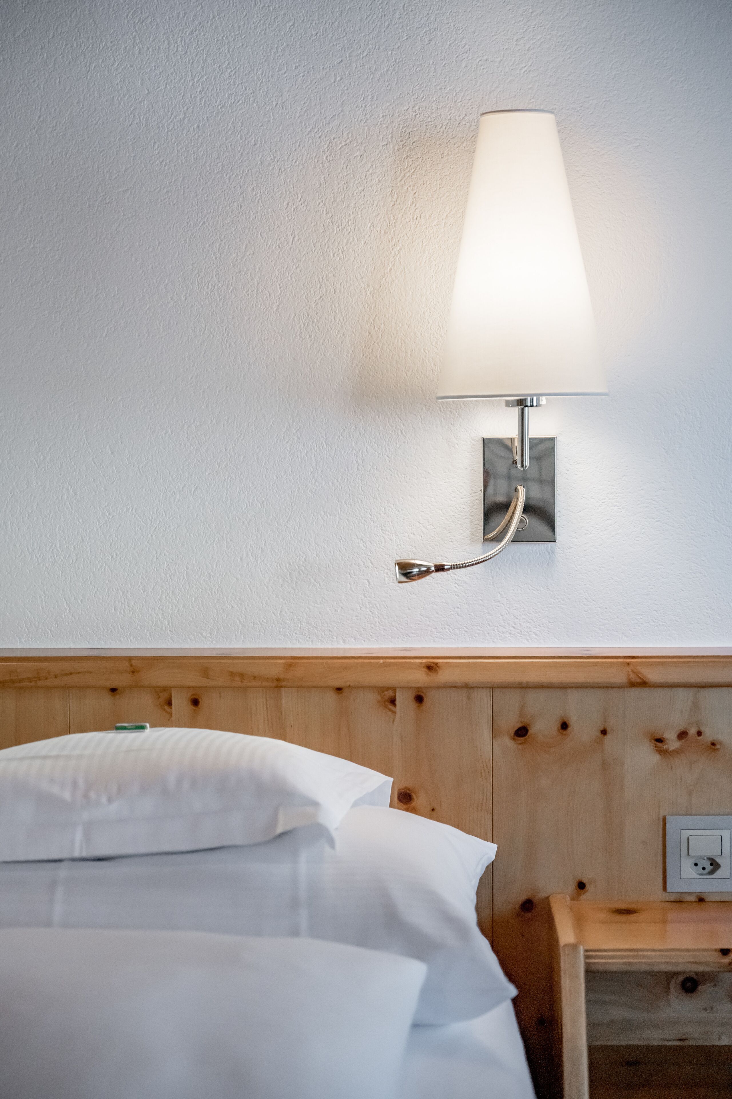 Habitación de hotel con cabecera de madera, cama blanca con almohadas, mesita de noche con lámpara y toma de corriente.
