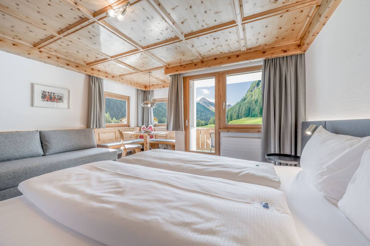 Ein Hotelzimmer mit Holzbalkendecke, einer Sitzecke, einem Bett mit weißer Bettwäsche, einem Schreibtisch und Blick auf Berge durch große Fenster.
