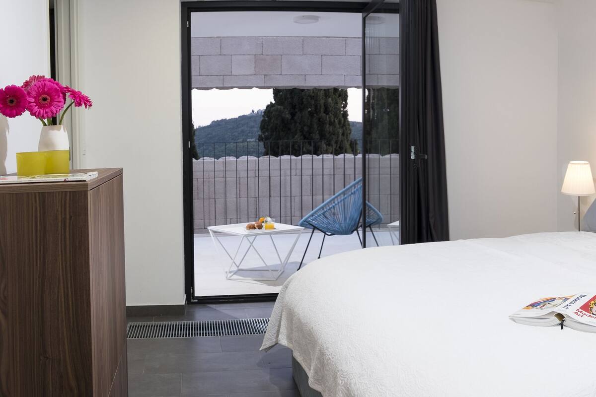 A hotel room with a bed, bedside lamps, a magazine on the bed, a desk with a vase of flowers, and a view of the outdoors through a glass door.