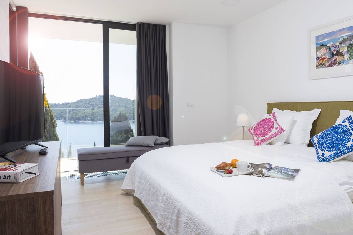 A hotel room with a bed, a desk, a TV, and a view of the sea.