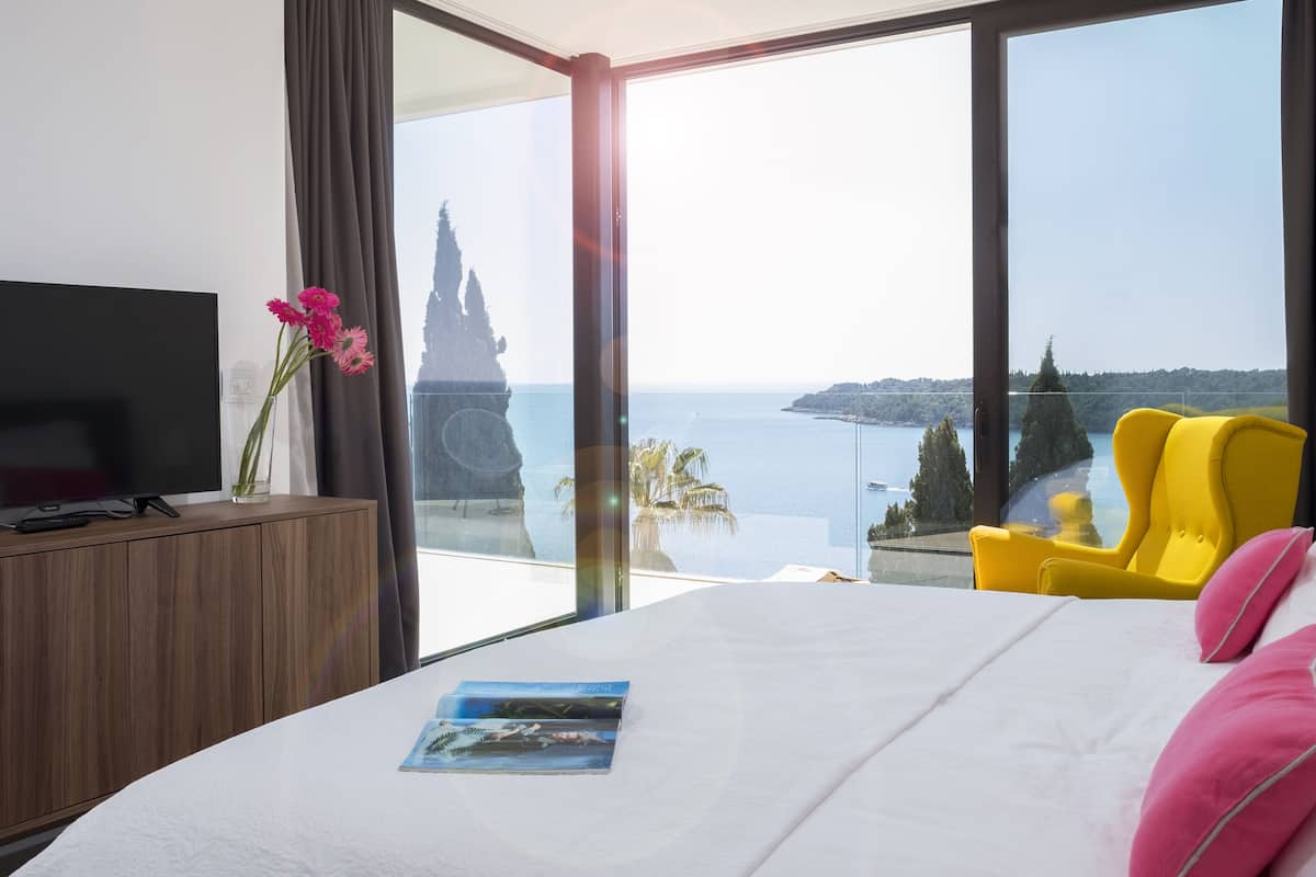 A modern hotel room with a large bed, a TV on a wooden cabinet, a yellow armchair, and a view of the sea and cliffs.