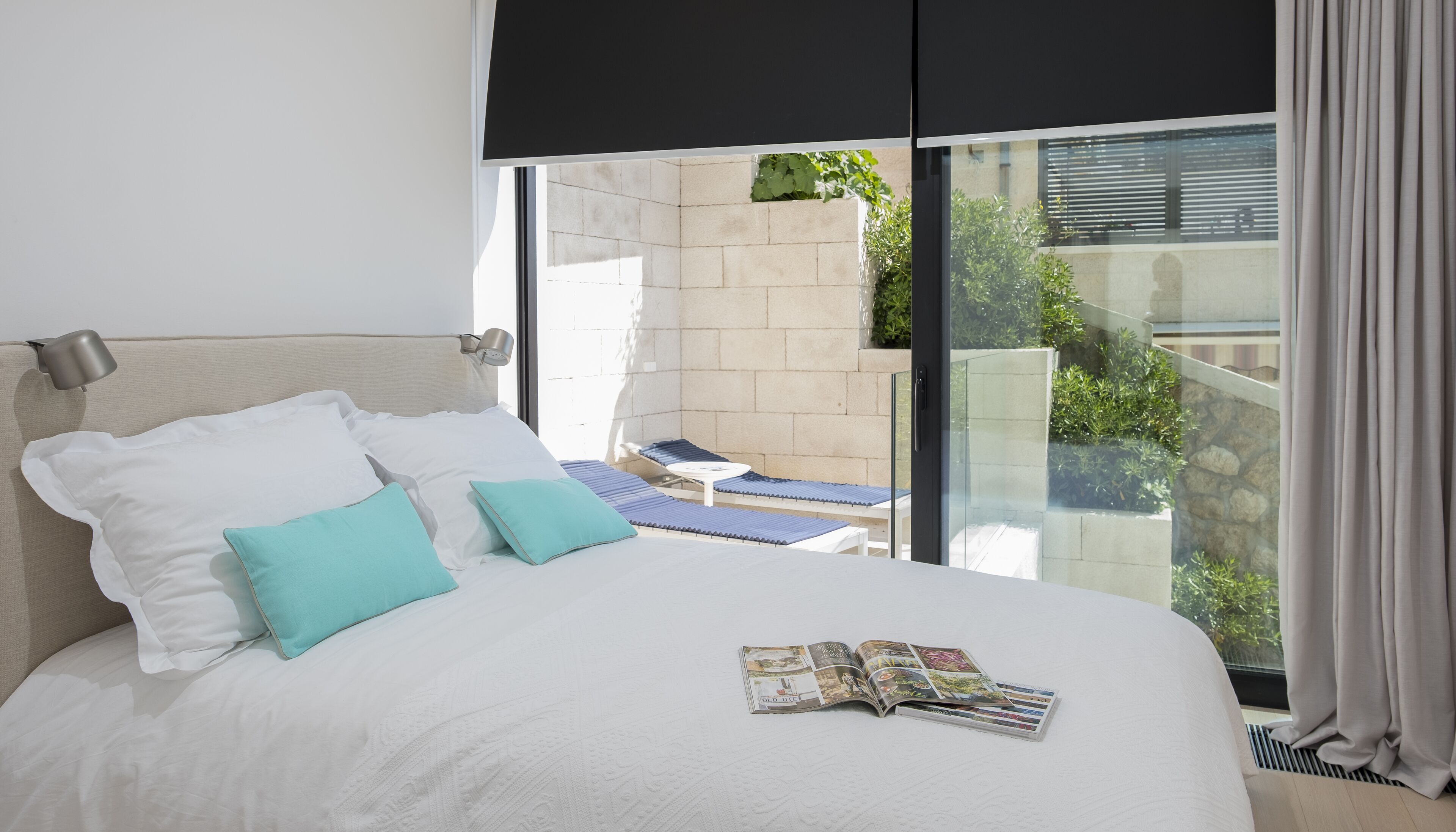 A hotel room with a bed, a magazine, a window with a view of greenery, and a balcony with a chair.