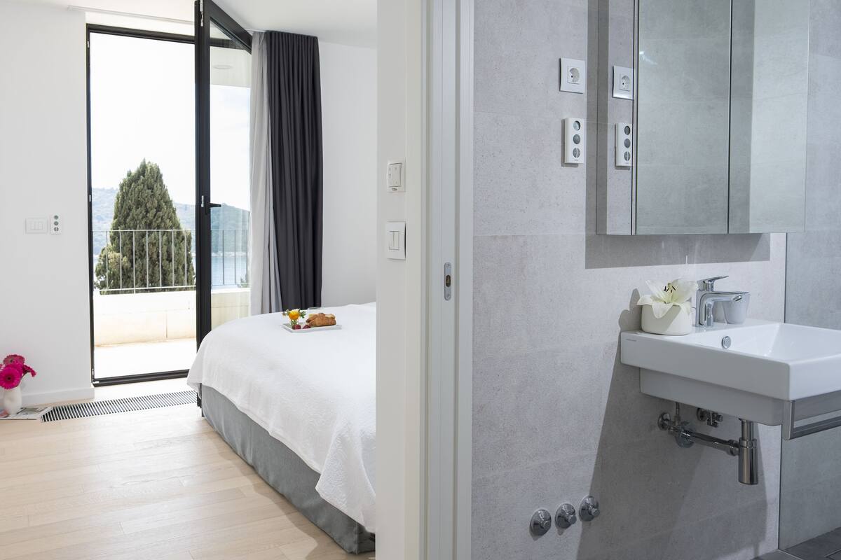 A modern hotel room with a bed, a bathroom with a sink and mirror, and a balcony with a view of the sea and trees.