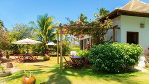 A garden with a wooden pergola, tables, and chairs, surrounded by lush greenery and flowering plants. - Pousada San Giovanni (Japaratinga)