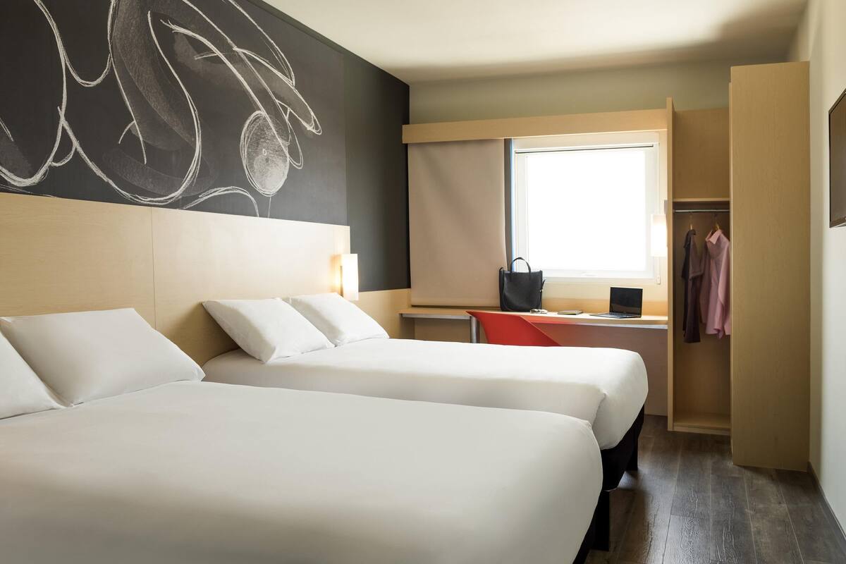 A hotel room with two beds, a desk, a chair, a window with curtains, and a wardrobe.