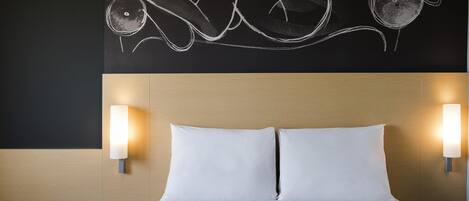 Una cama con sábanas blancas, dos lámparas de noche y una pared con un dibujo en la pizarra.