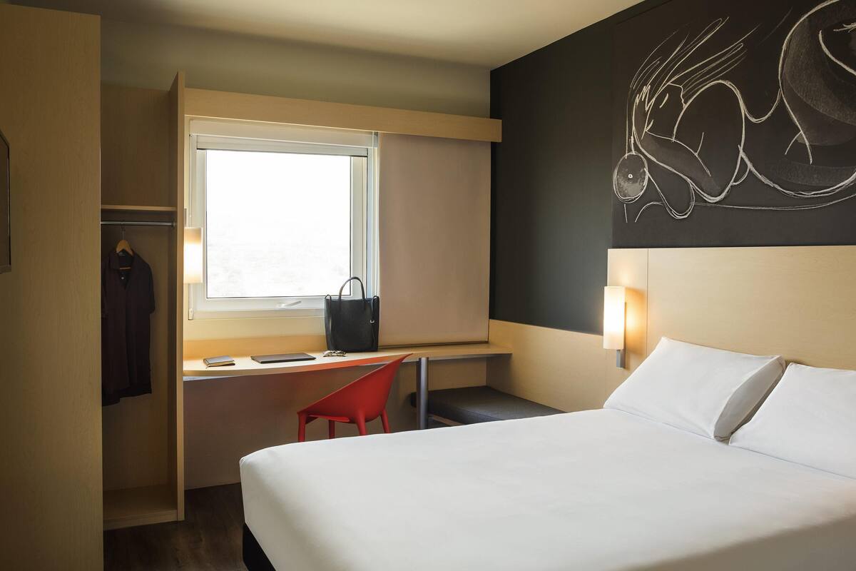 A hotel room with a bed, a desk, a chair, a wardrobe, and a wall art piece.