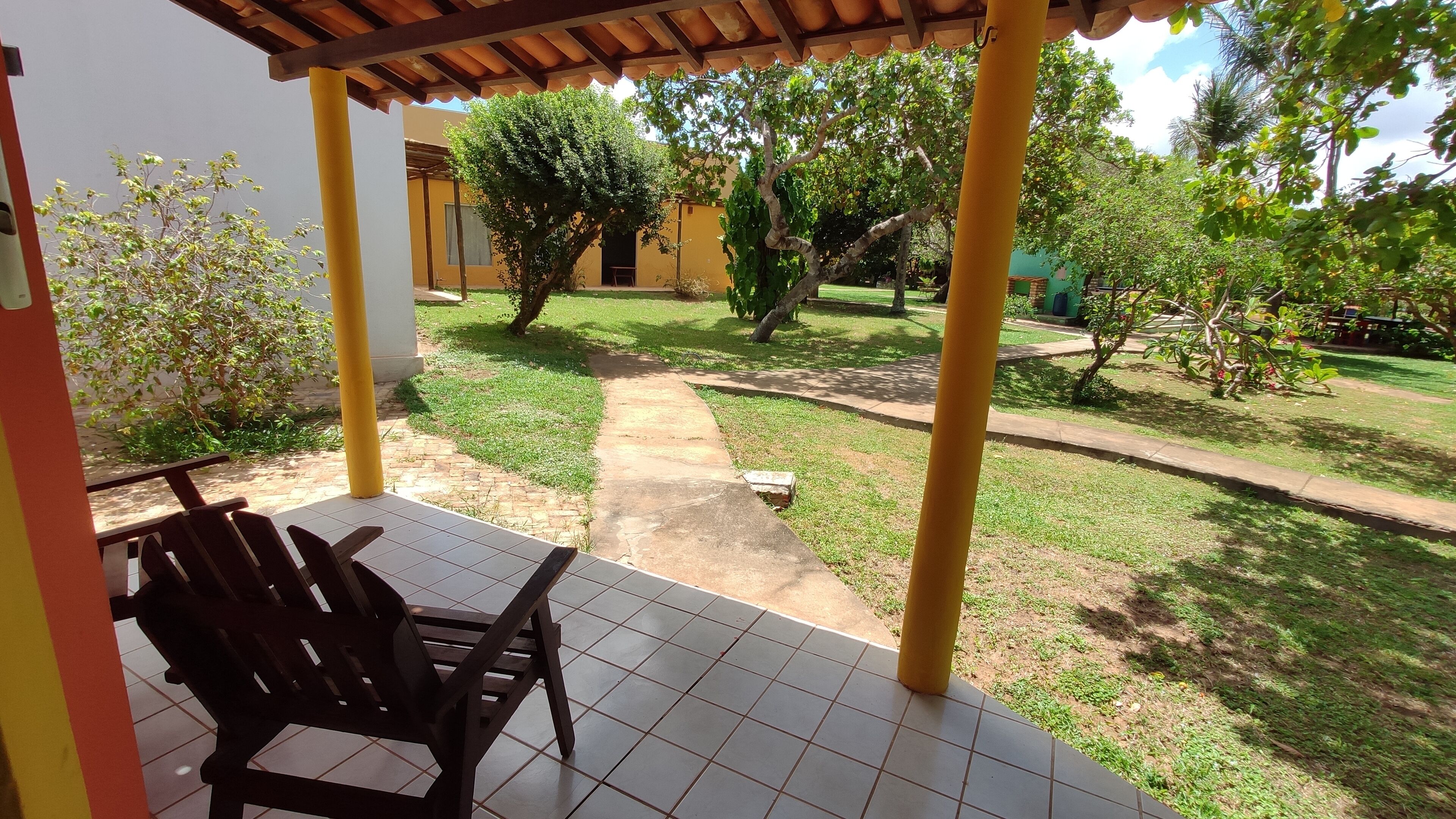 A covered patio area with a tiled floor, a wooden bench, and a pathway leading to a grassy area with trees.