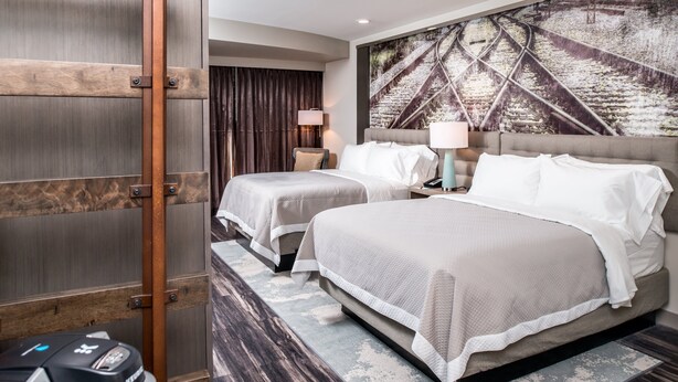 A hotel room with two beds, a large window with curtains, a wall-mounted artwork, and a small table with a lamp.