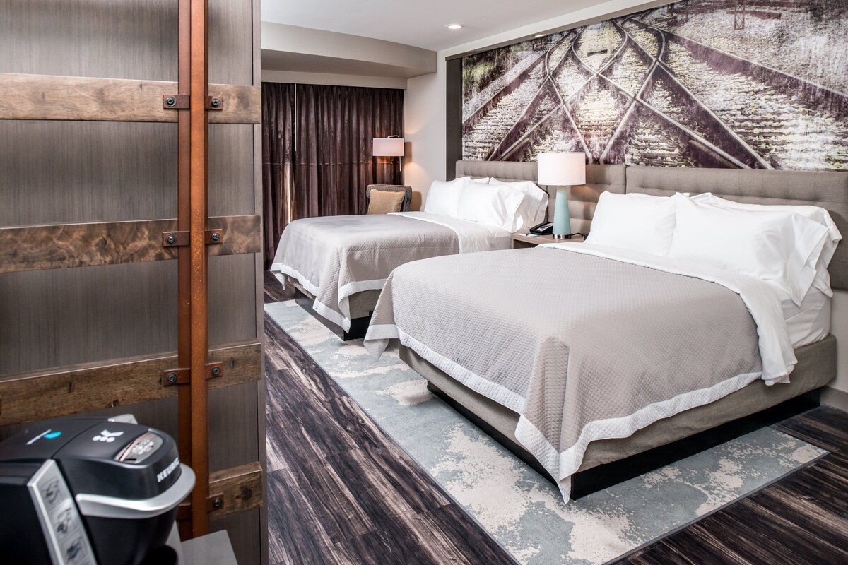 Une chambre d’hôtel avec deux lits, une grande fenêtre avec des rideaux, une œuvre d’art accrochée au mur et une petite table avec une lampe.