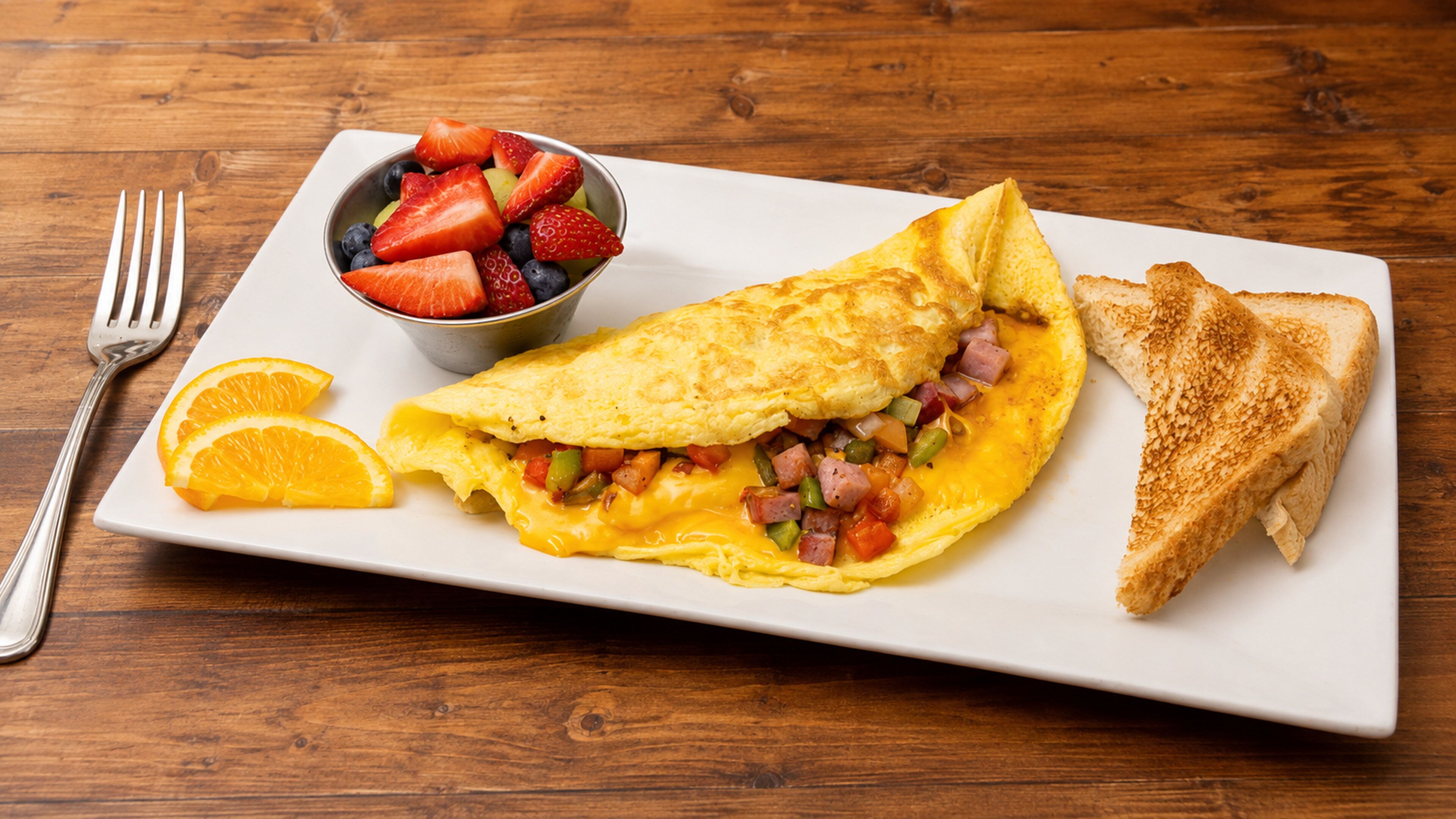 A breakfast plate with an omelette, toast, and a bowl of fruit.