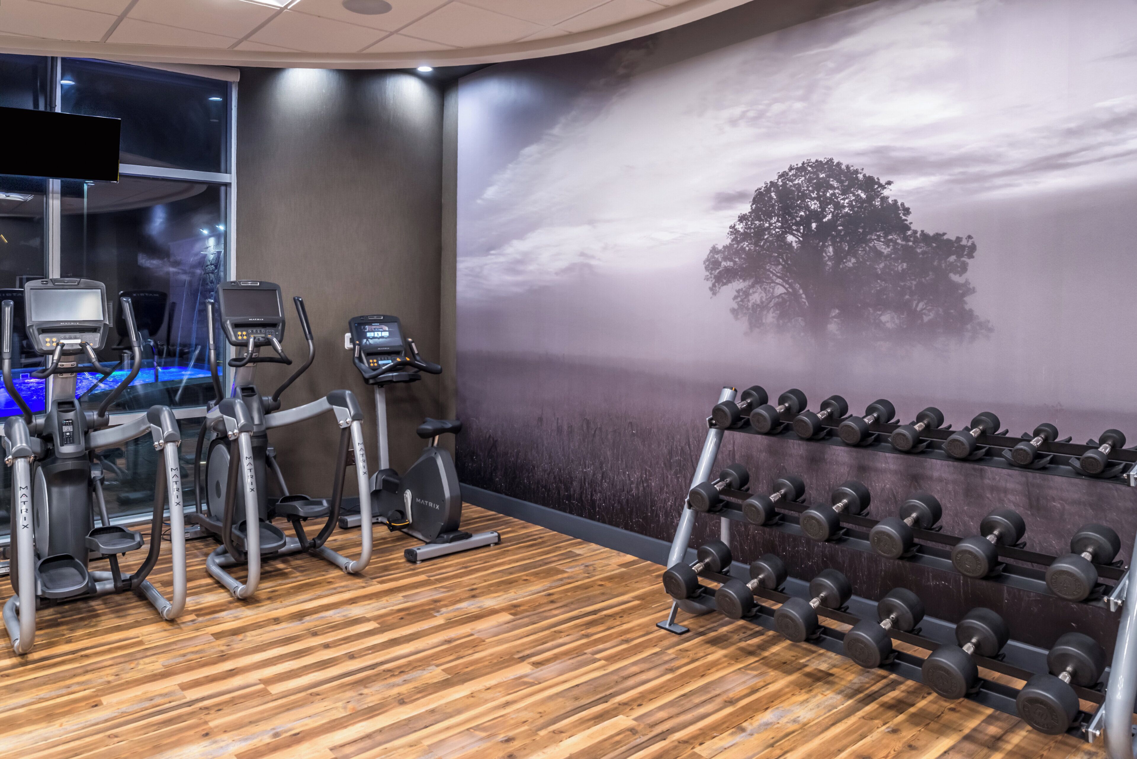 Un gimnasio con equipo de ejercicio, un televisor montado en la pared y un ventanal grande.