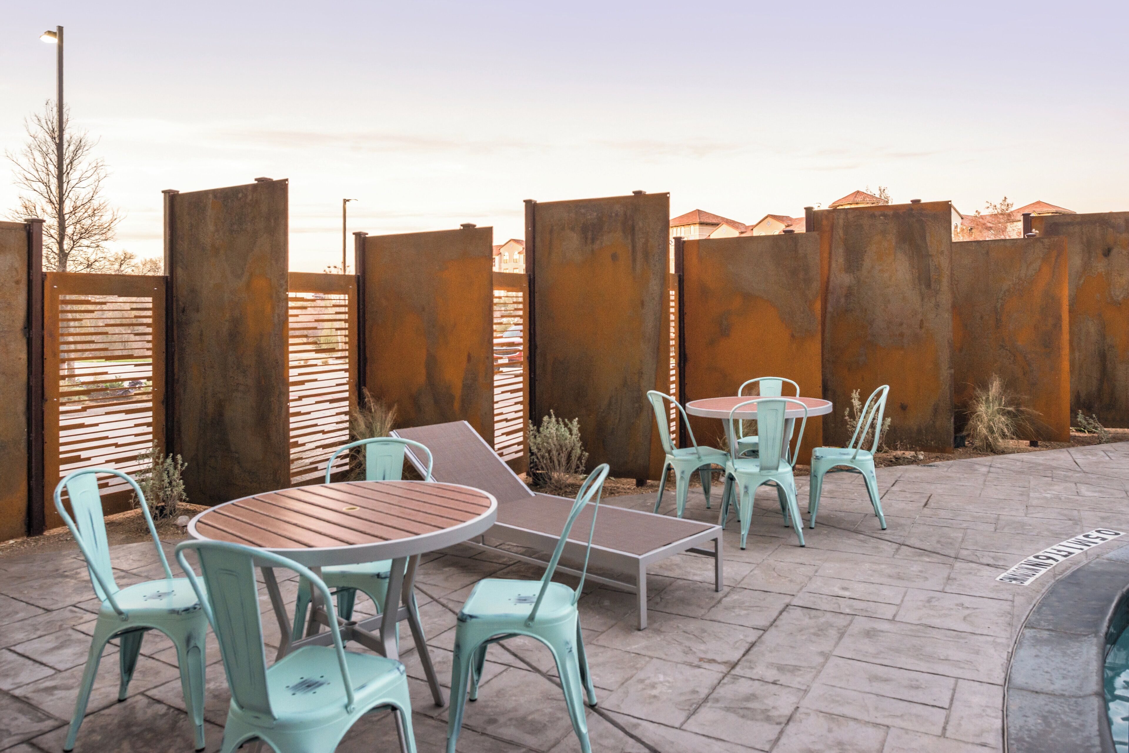 Un patio al aire libre con sillas y mesas de metal, un banco de madera y una pared de ladrillo.