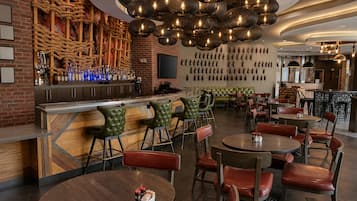 A bar area with a brick wall, wooden bar counter, and a variety of bottles. There are multiple seating areas with leather chairs and tables.