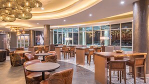 A modern hotel lobby with round tables, leather chairs, and a large window offering a view of the city.