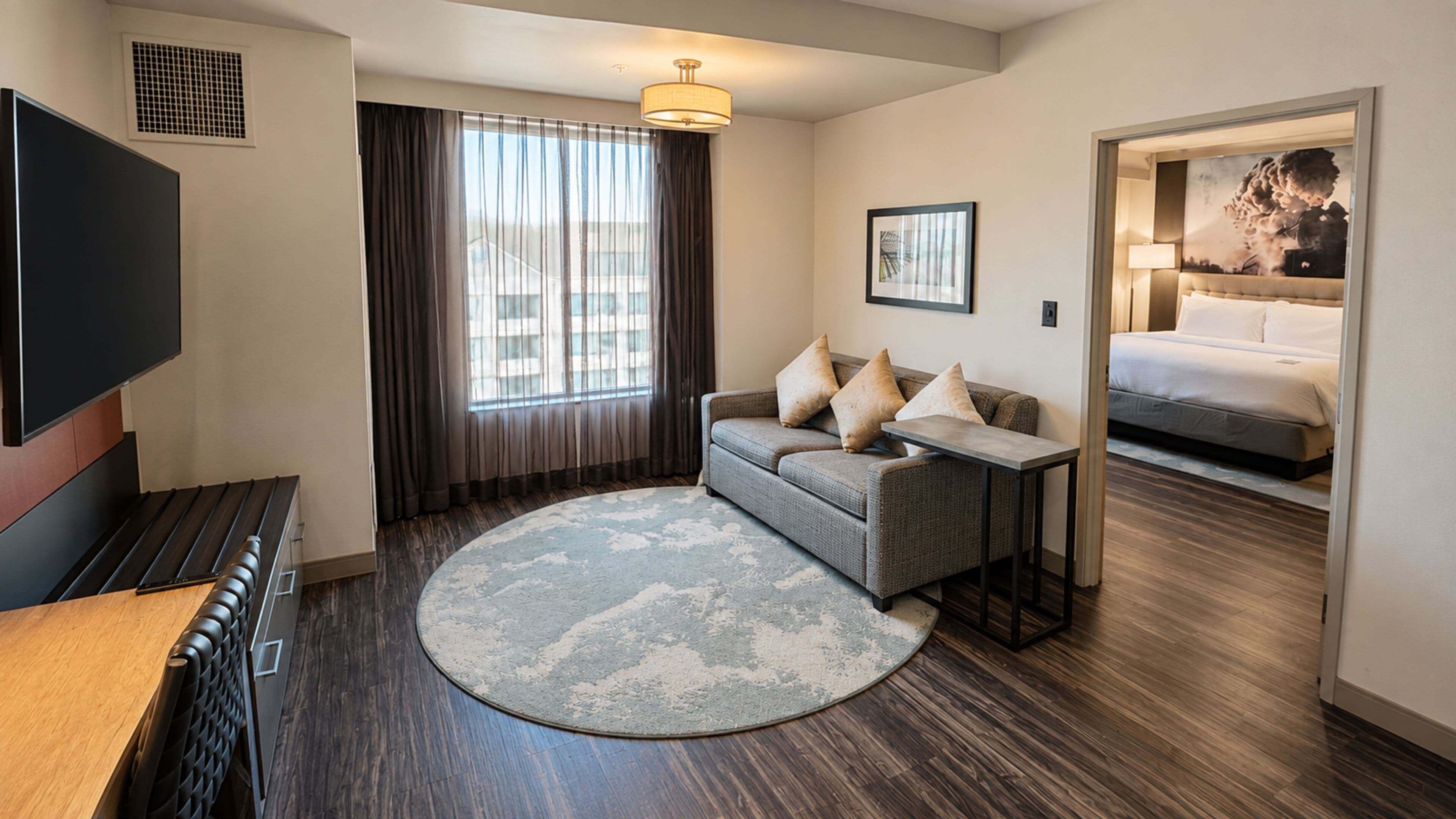 A hotel room with a flat-screen TV, a sofa, a small table, a round rug, and a bed with a headboard.