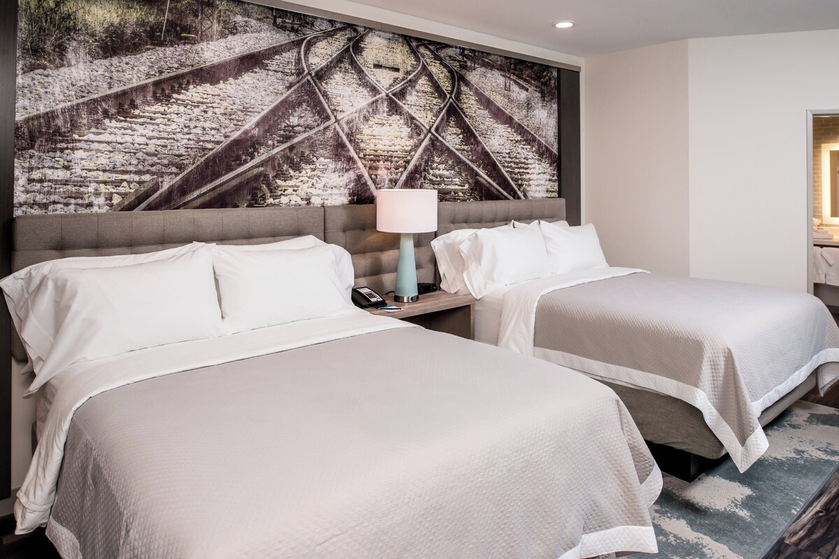 Une chambre d’hôtel avec deux lits, une grande fresque murale, une table de chevet avec une lampe, et une vue sur l’intérieur de la pièce.