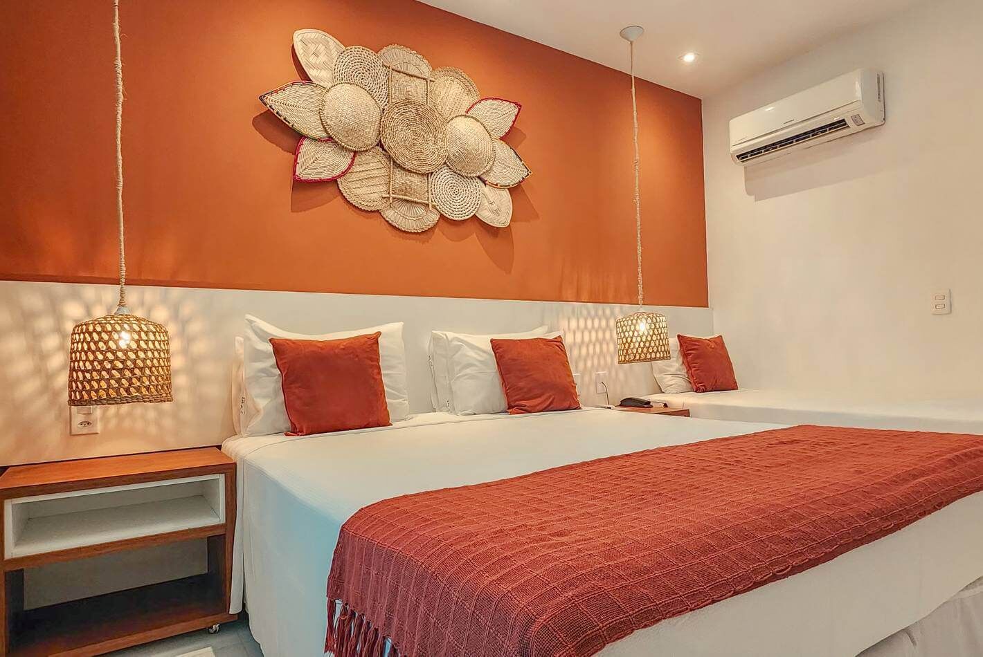 Un dormitorio moderno con una cama grande, ropa de cama blanca y un plaid rojo. Hay dos mesitas de noche con lámparas, y un aire acondicionado instalado en la pared.