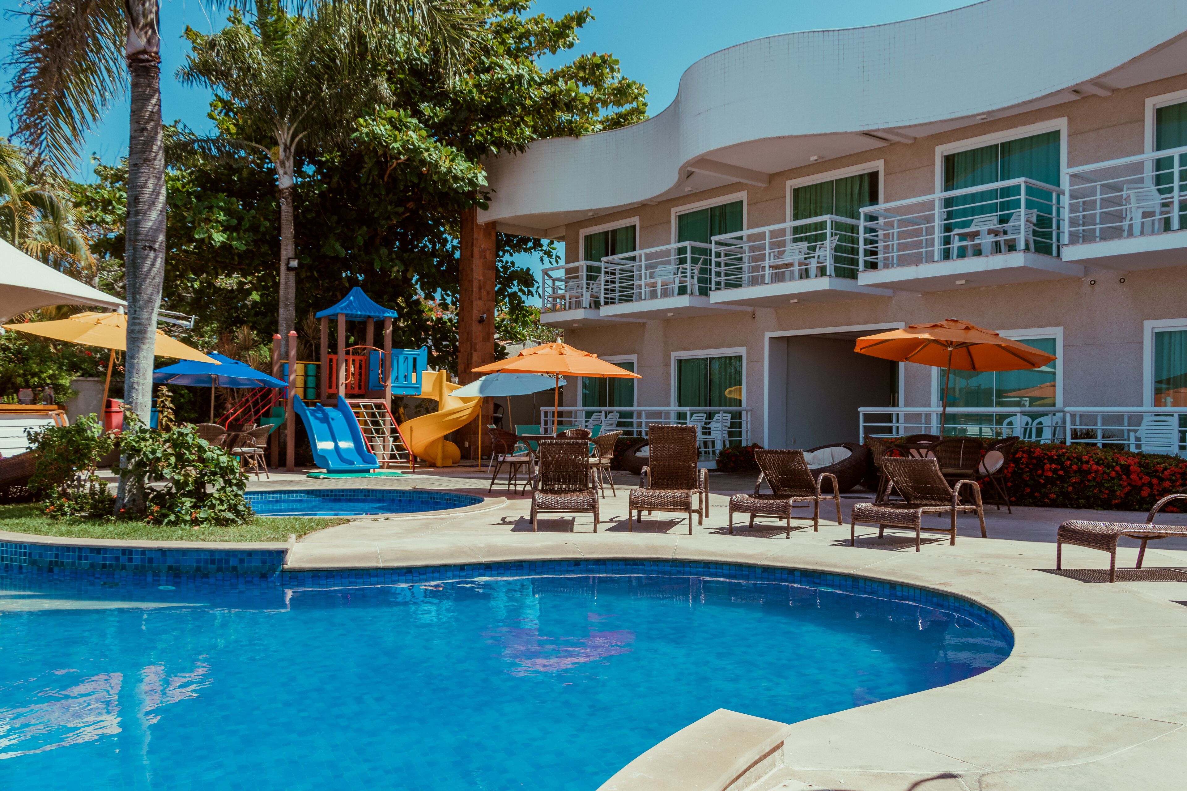 A swimming pool with lounge chairs and umbrellas, a children's play area, and a multi-story building with balconies.