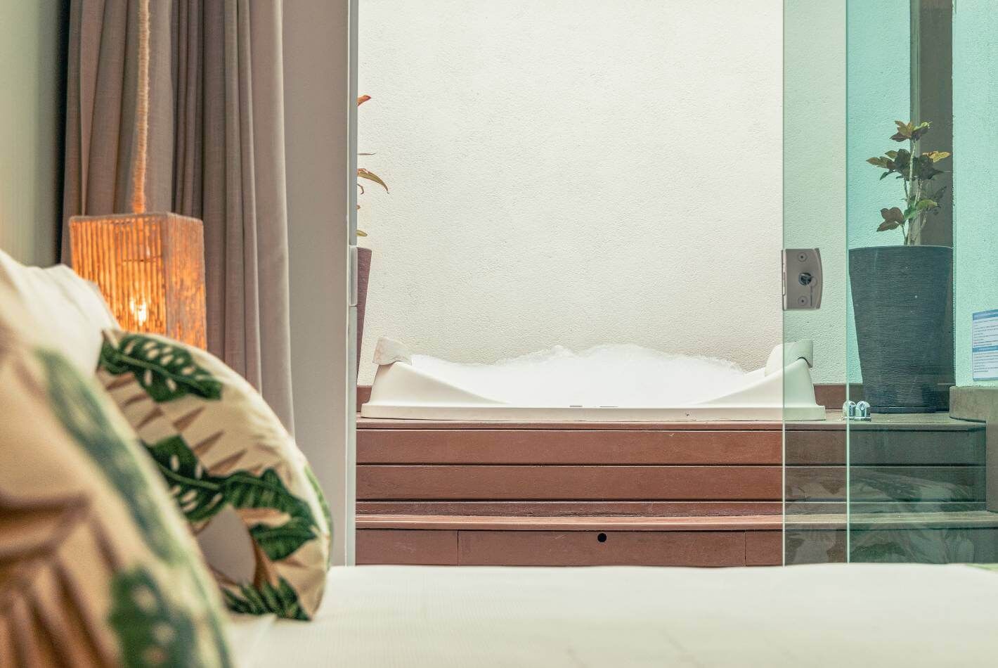 Habitación de hotel con una cama, una ventana con vista a una pared blanca, una mesita de noche de madera y una lámpara.
