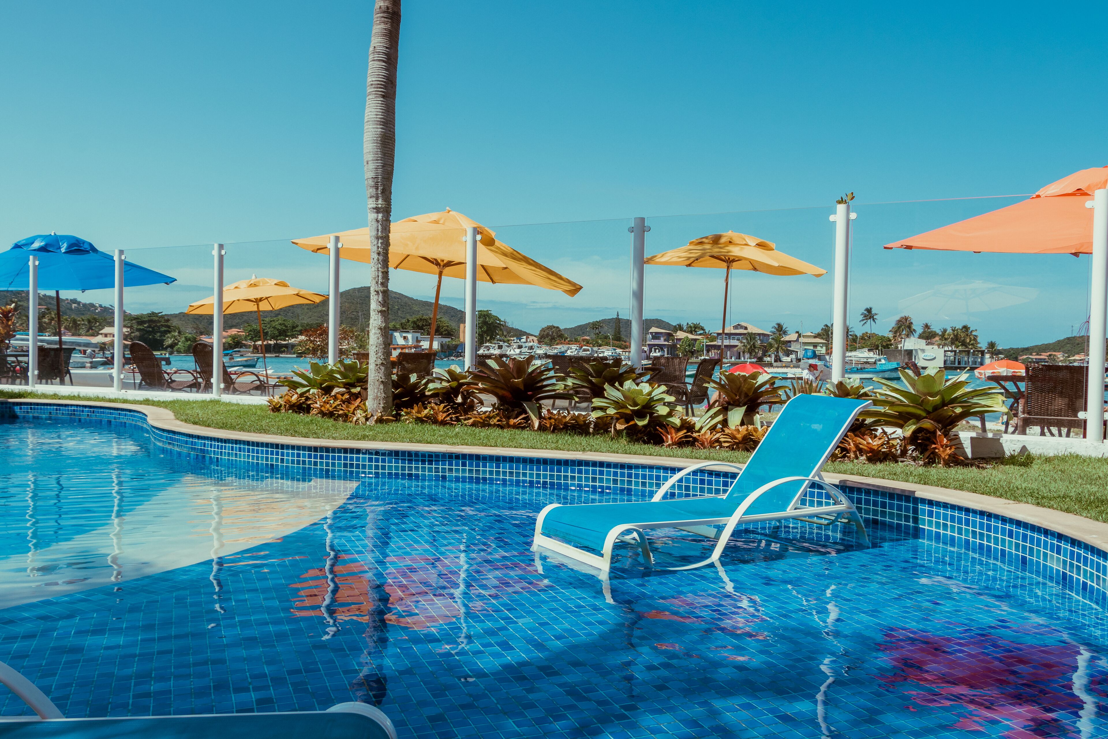 A swimming pool with tiled blue edges, surrounded by lounge chairs and umbrellas, with a view of hills in the background.