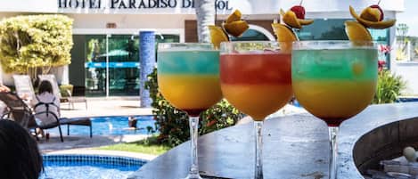 Three colorful cocktails with fruit garnishes on a bar counter, a pool, and a hotel named "Hotel Paradiso" in the background.