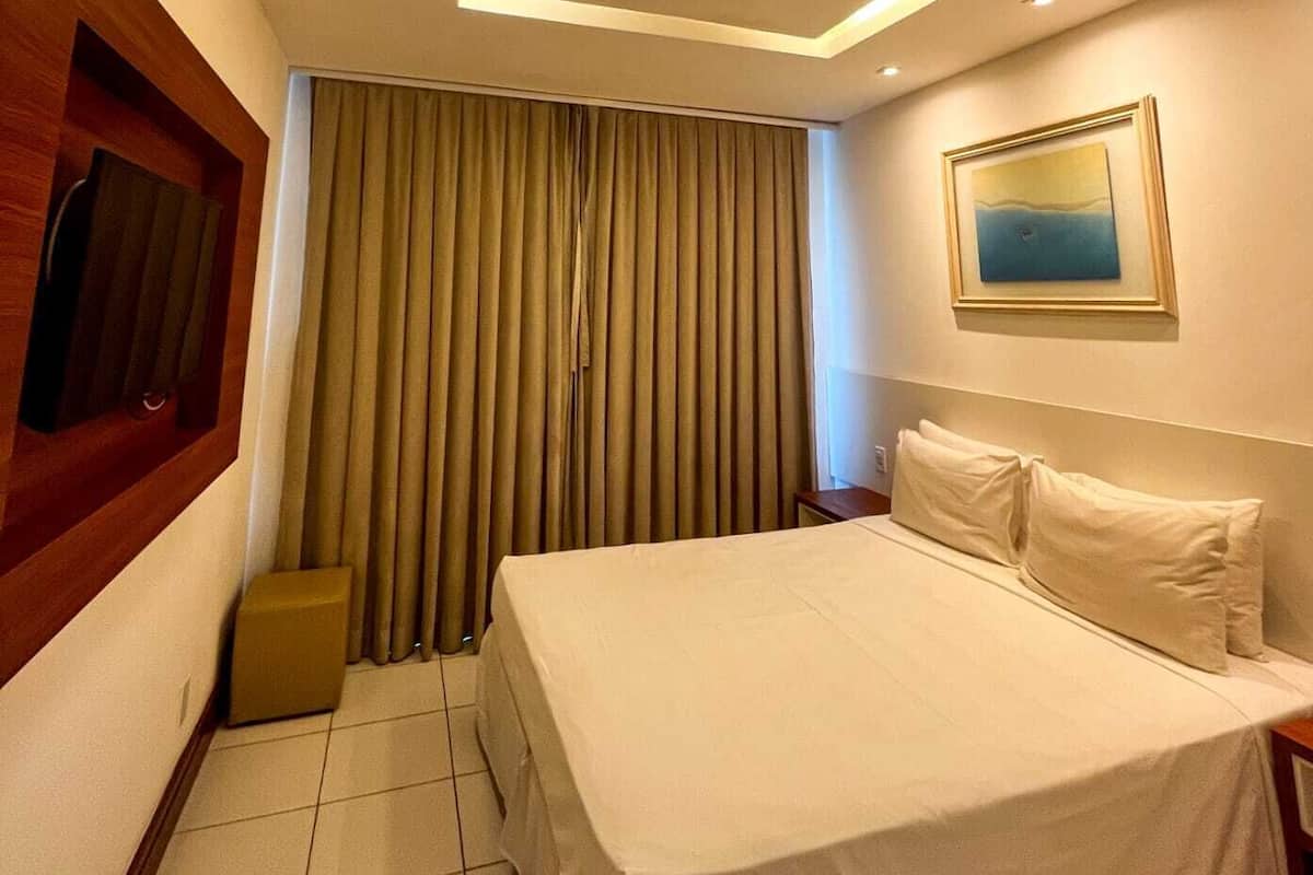 Una habitación de hotel con cama, cortinas, un cuadro y una lámpara empotrada en el techo.