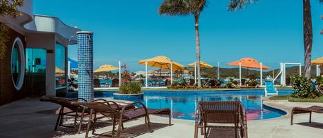 A pool area with lounge chairs and umbrellas, surrounded by palm trees and a clear blue sky.