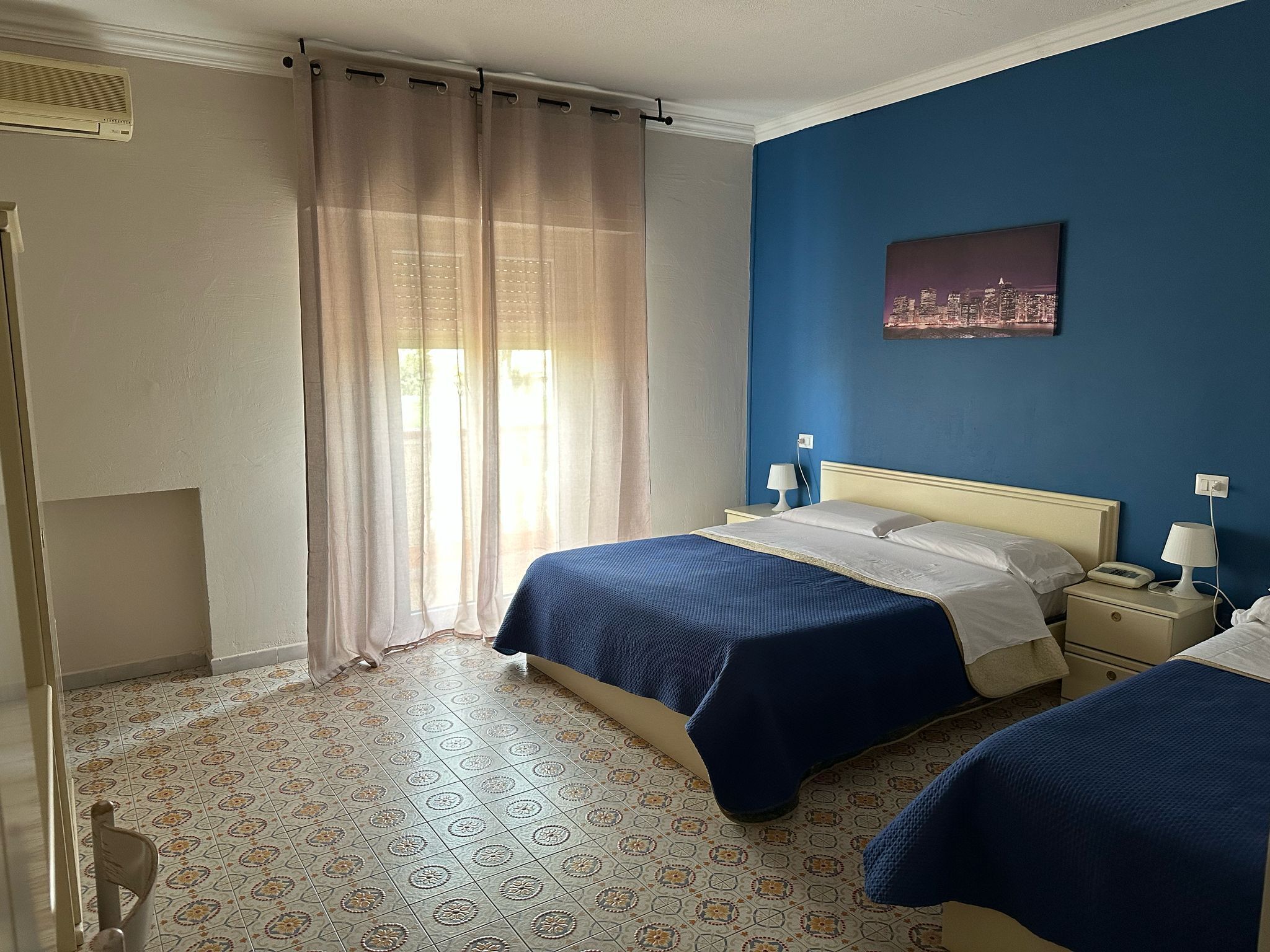 A hotel room with two beds, a cityscape painting, a window with sheer curtains, and patterned floor tiles.