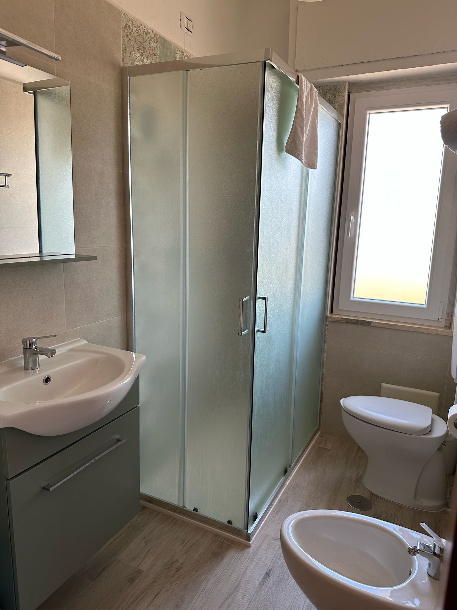 A bathroom with a glass shower enclosure, a sink, a toilet, and a window providing natural light.