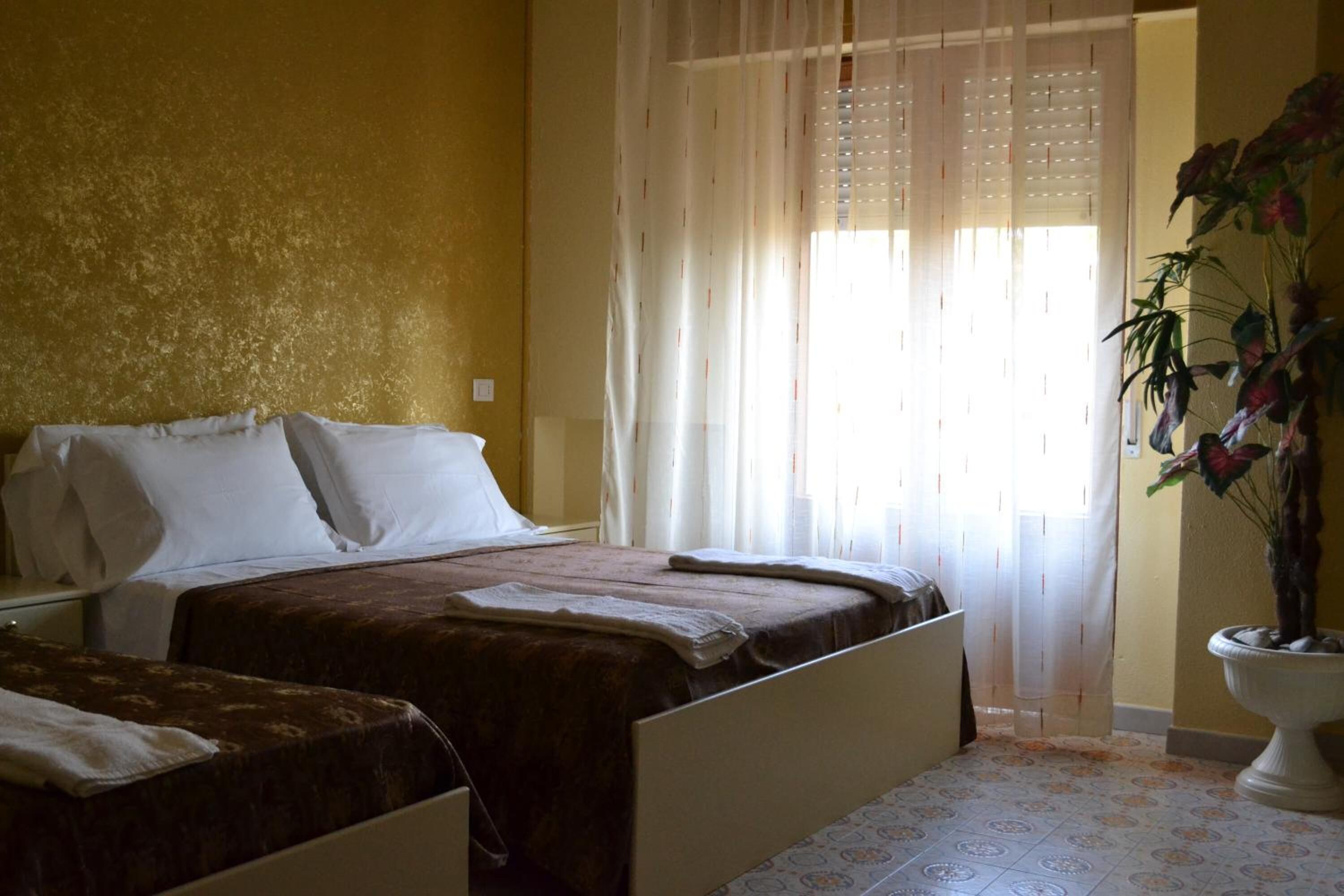A bedroom with two beds, a large window with sheer curtains, and a potted plant.