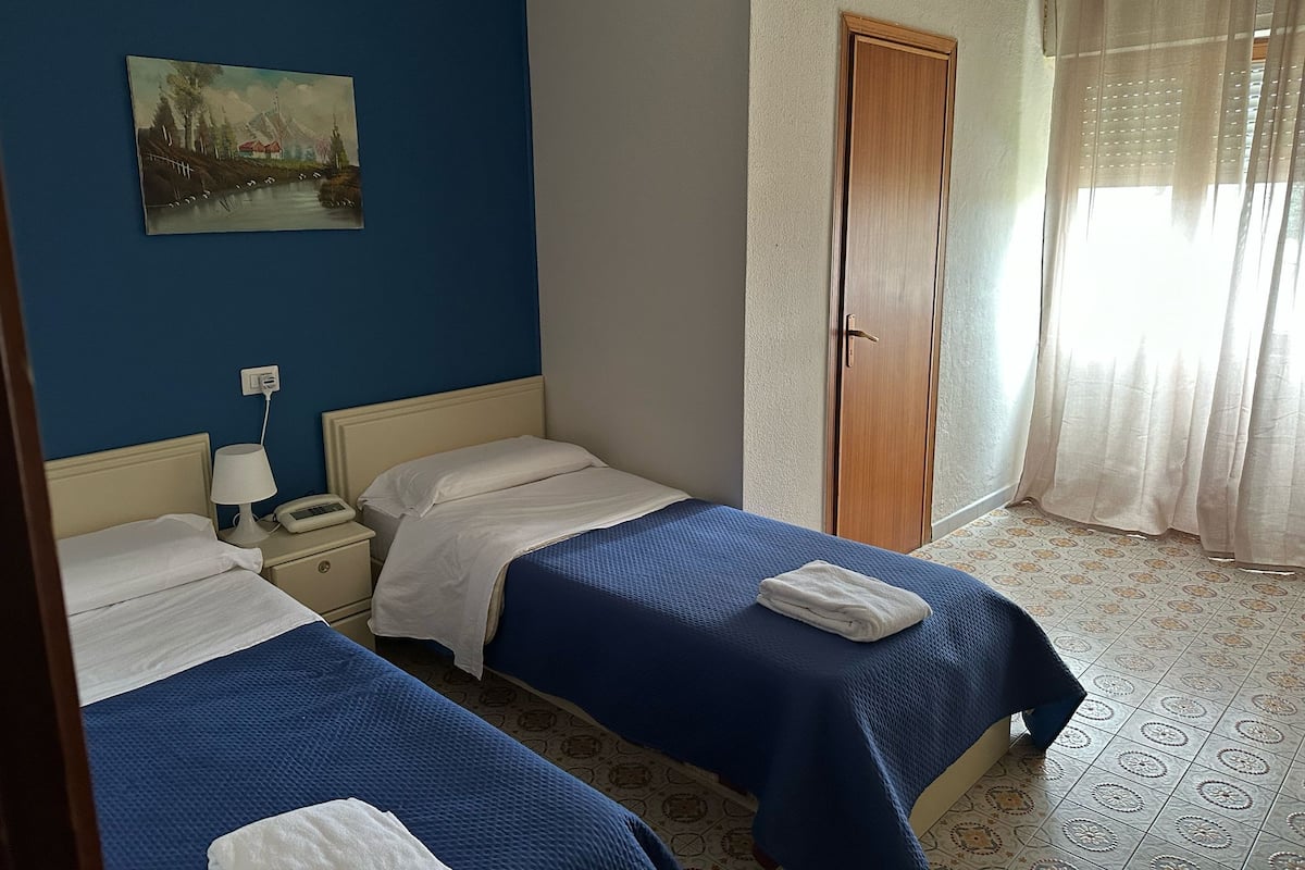Une chambre d’hôtel avec deux lits, une table de chevet avec une lampe, un tableau au mur et une porte avec un rideau.