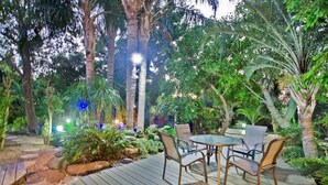 An outdoor patio area with a round table and chairs, surrounded by lush greenery and palm trees. - Siesta (Ramot)