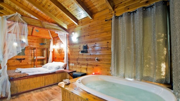 A wooden cabin room with a bed, a small table, a bathtub, and a window with curtains. - Siesta (Ramot)