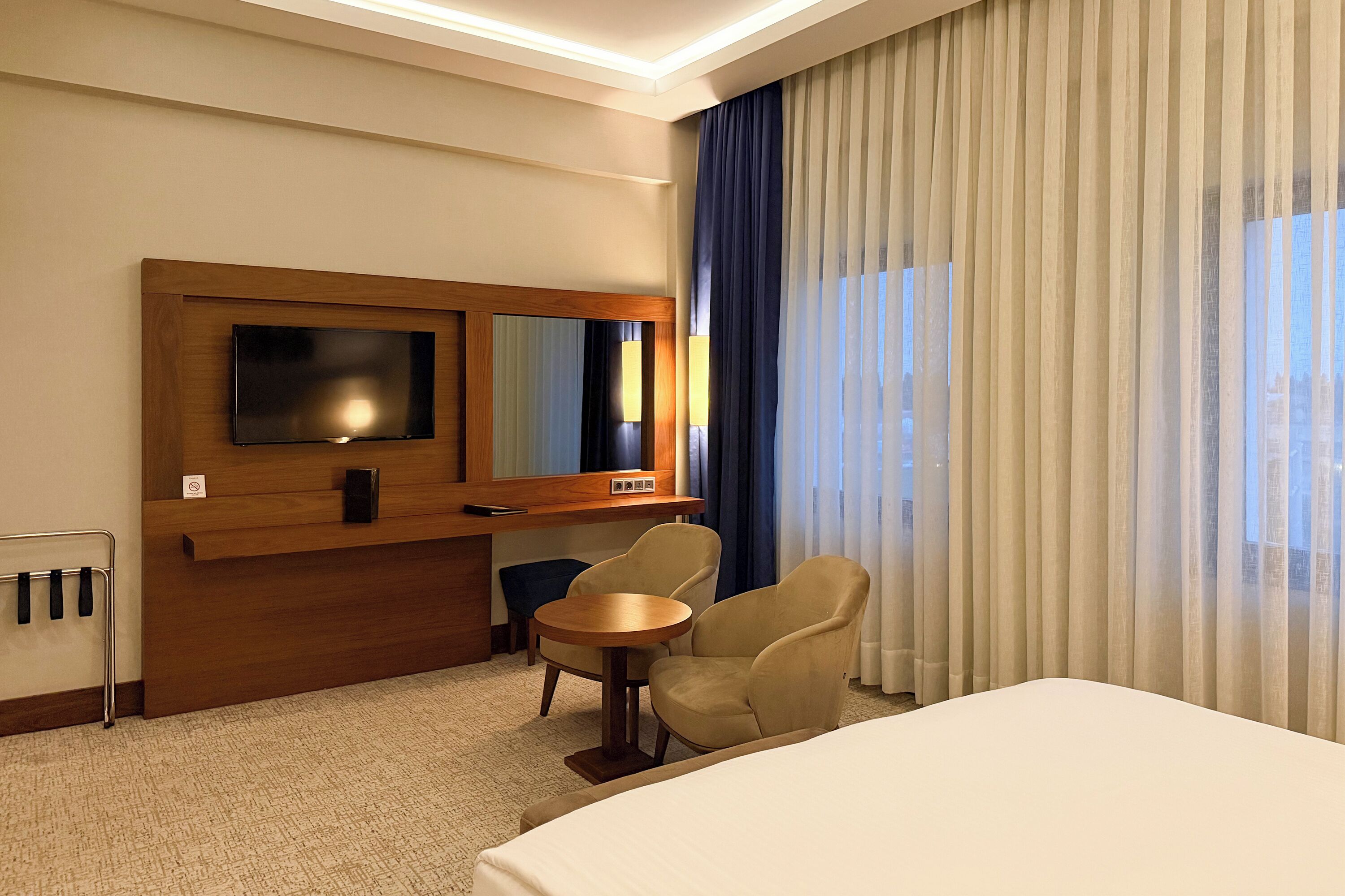 A hotel room with a bed, a TV, a desk, two chairs, and a large window with curtains.
