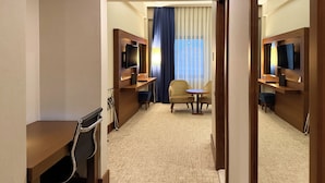 Habitación de hotel con escritorio, dos sillas, una mesita, televisión y una ventana grande con cortinas.
