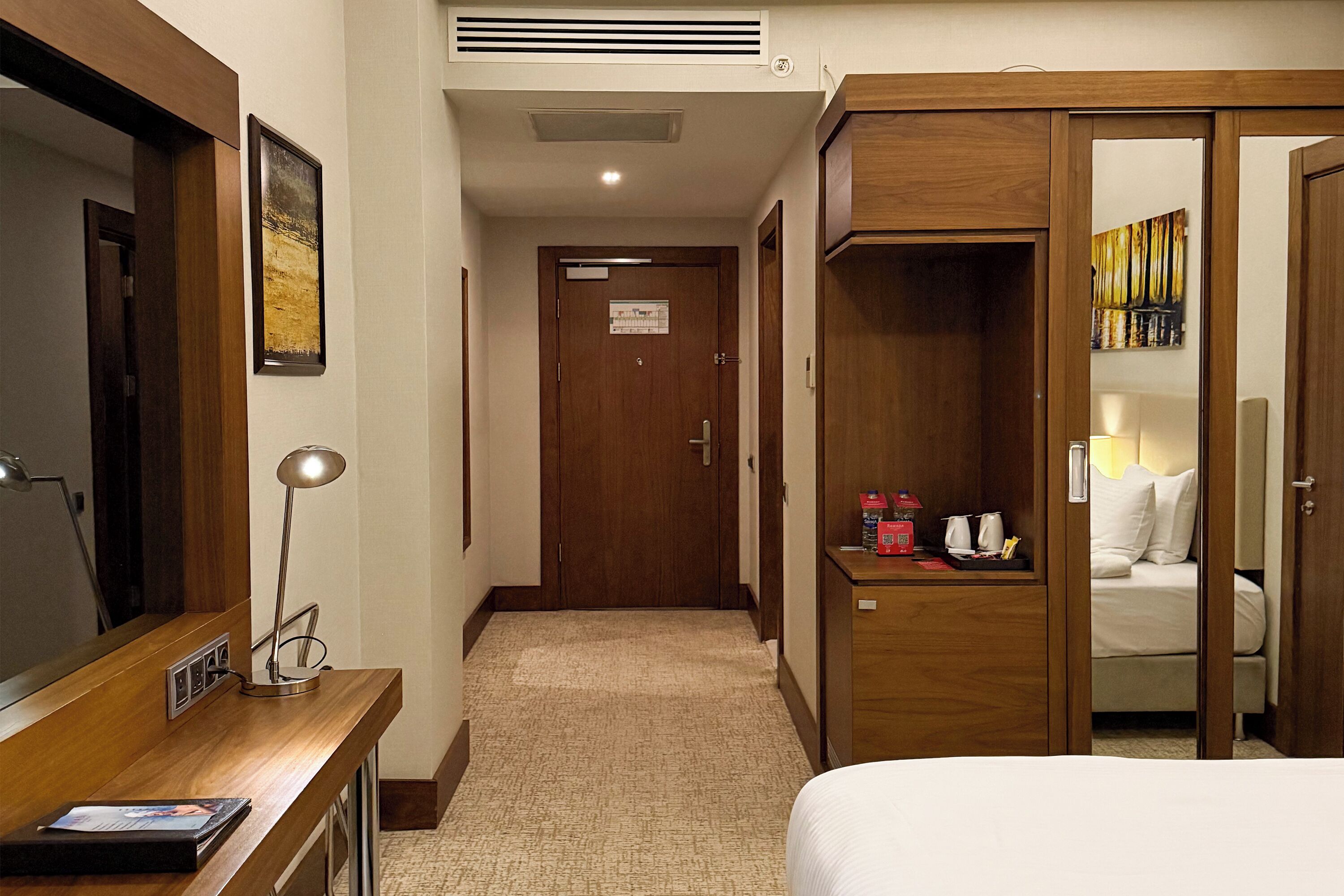 A hotel room with a bed, a desk with a lamp, a wardrobe, and a door leading to another room.