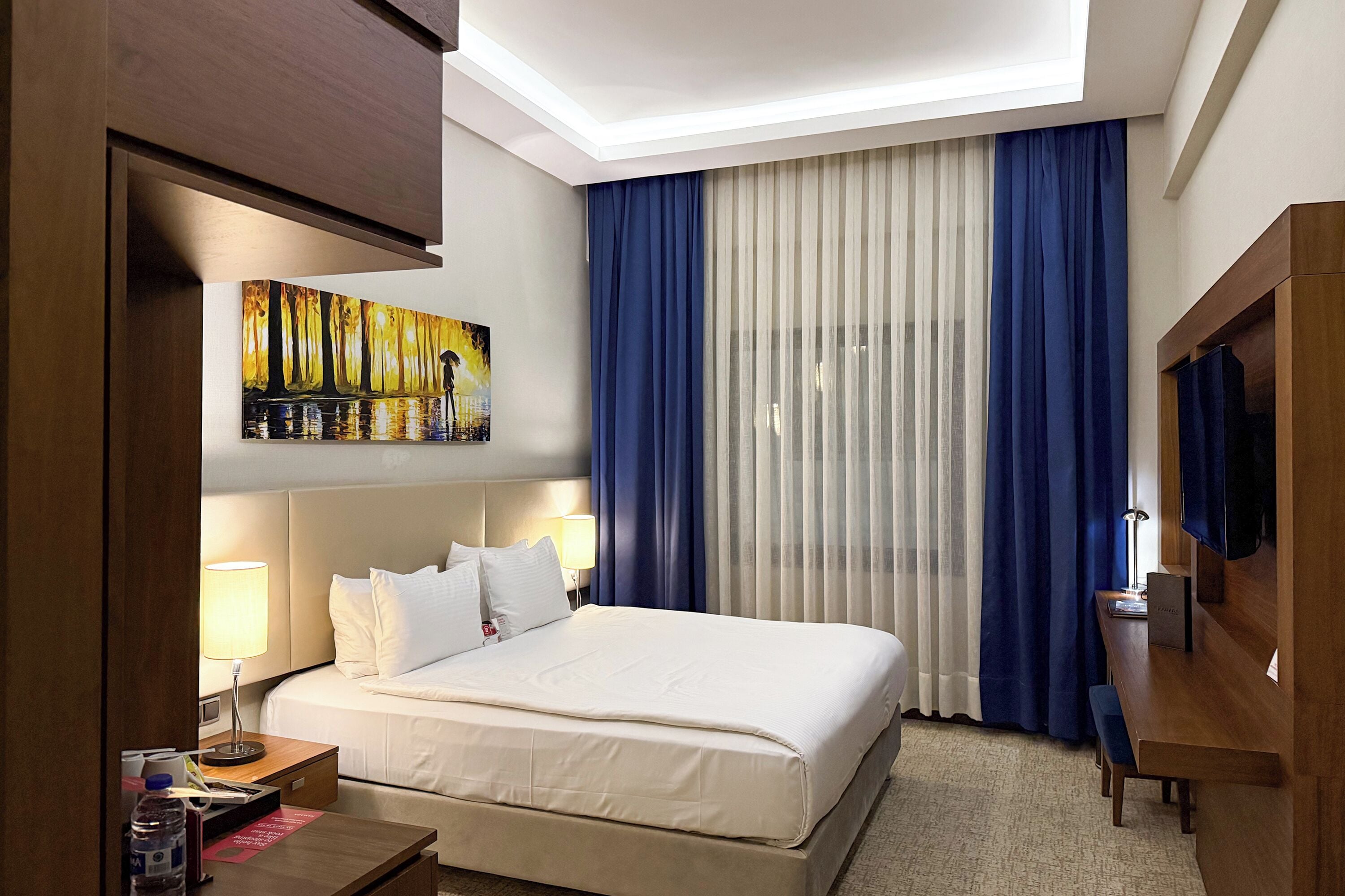 A hotel room with a large bed, a desk, a TV, and a painting on the wall.