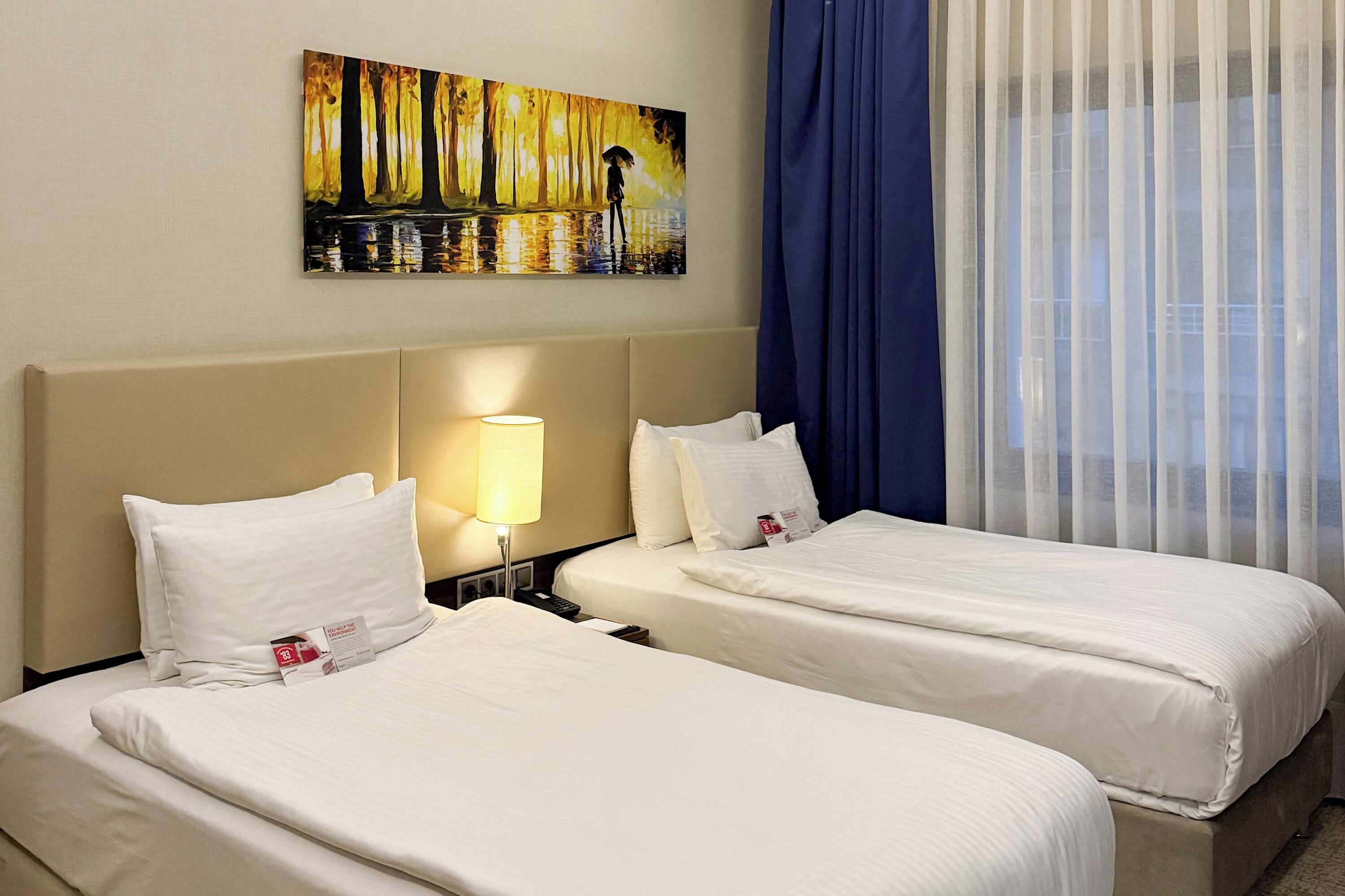 Une chambre d’hôtel avec deux lits, un tableau au mur, une lampe et une fenêtre avec des rideaux.