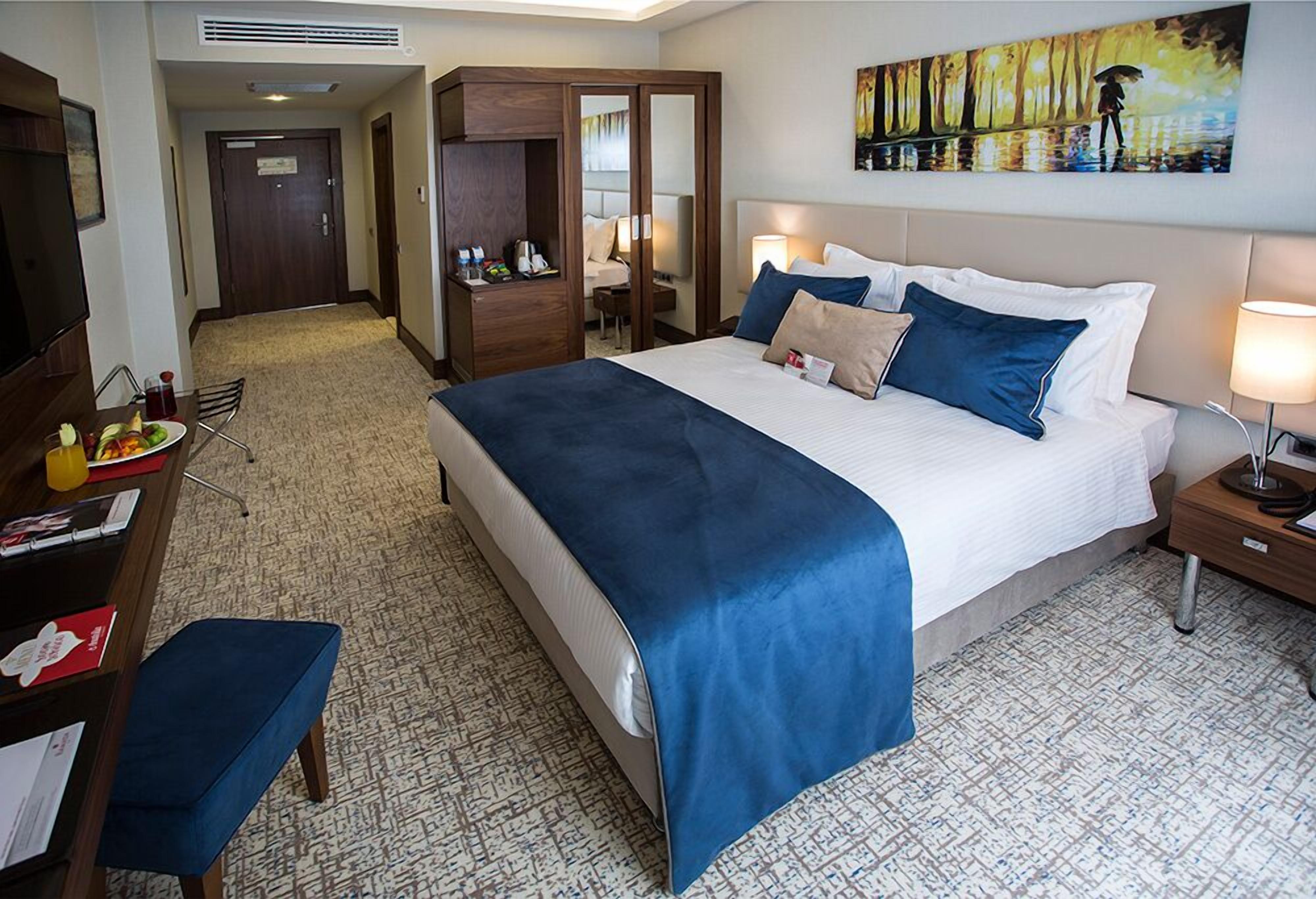 A hotel room with a large bed, a desk with a chair, a small table with a chair, a wardrobe, and a painting on the wall.