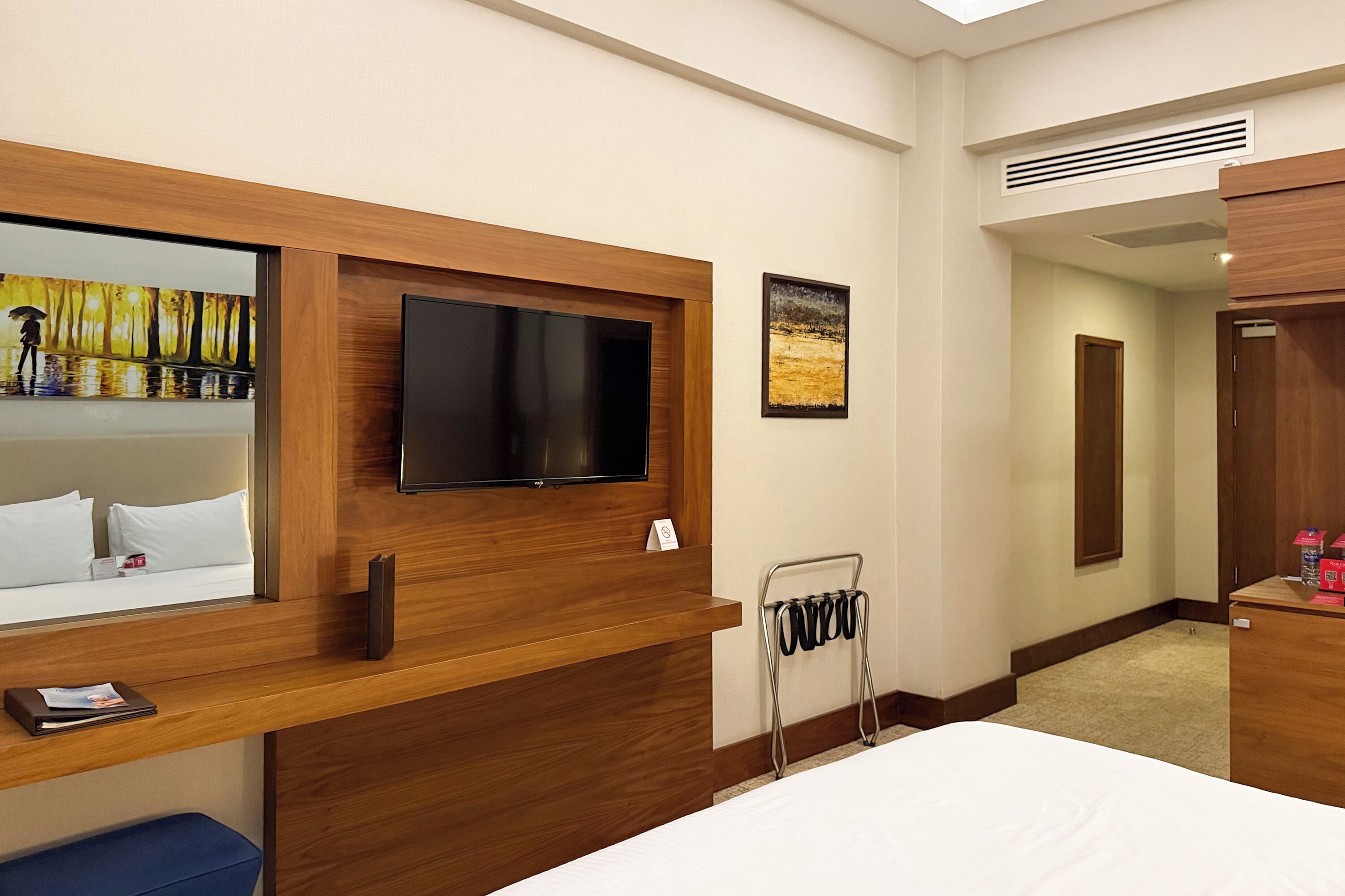 A hotel room with a wooden headboard, a flat-screen TV mounted on the wall, a small desk with a chair, a bedside table, and a bed with white bedding.