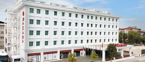 A white multi-story hotel with a flat facade, a covered entrance area, and a parking lot with a bus and a car.