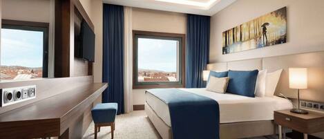 A hotel room with a bed, a desk, a chair, a TV, a painting, and a window with a city view.