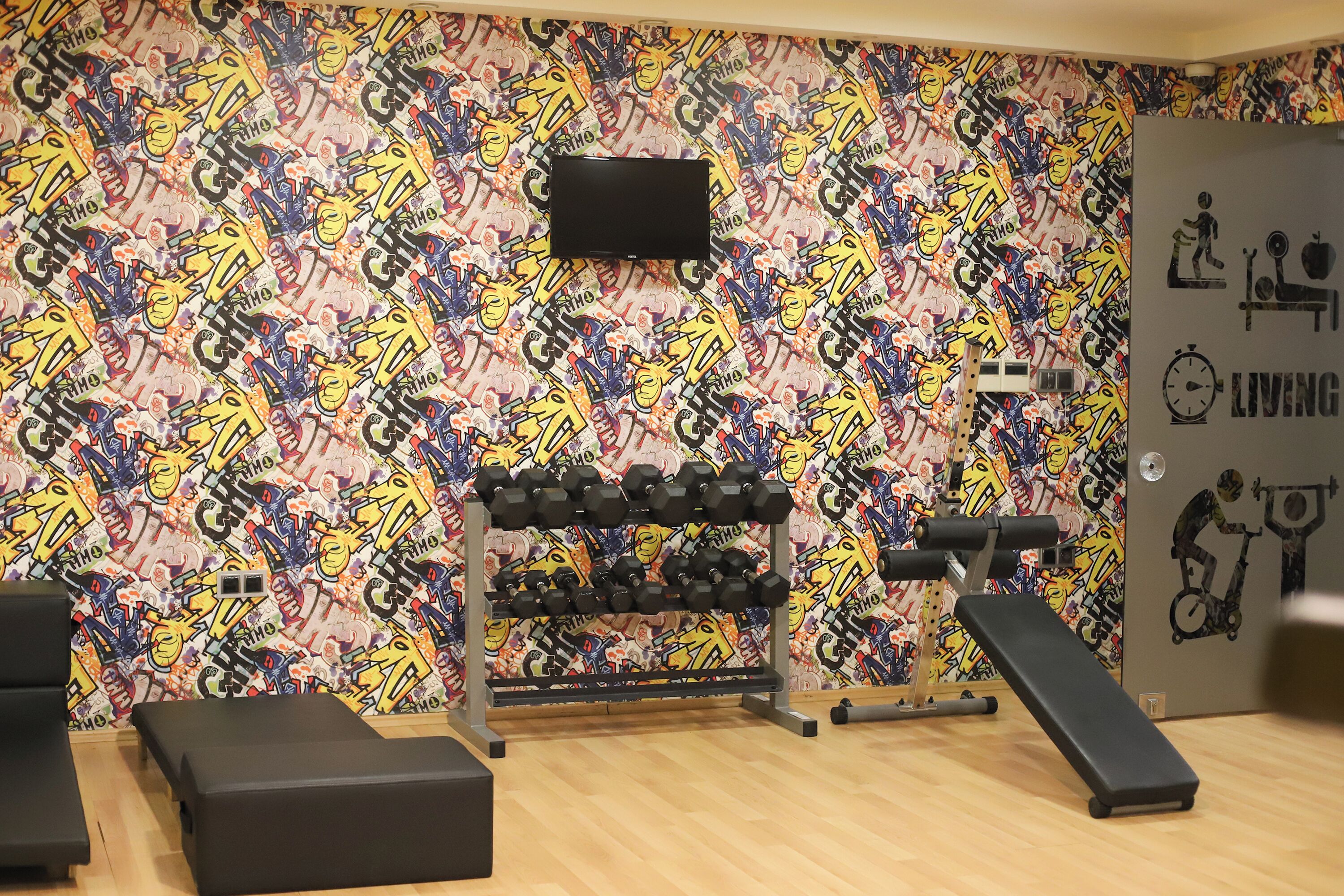 A gym with a colorful graffiti wall, a TV mounted on the wall, and various exercise equipment including dumbbells and benches.