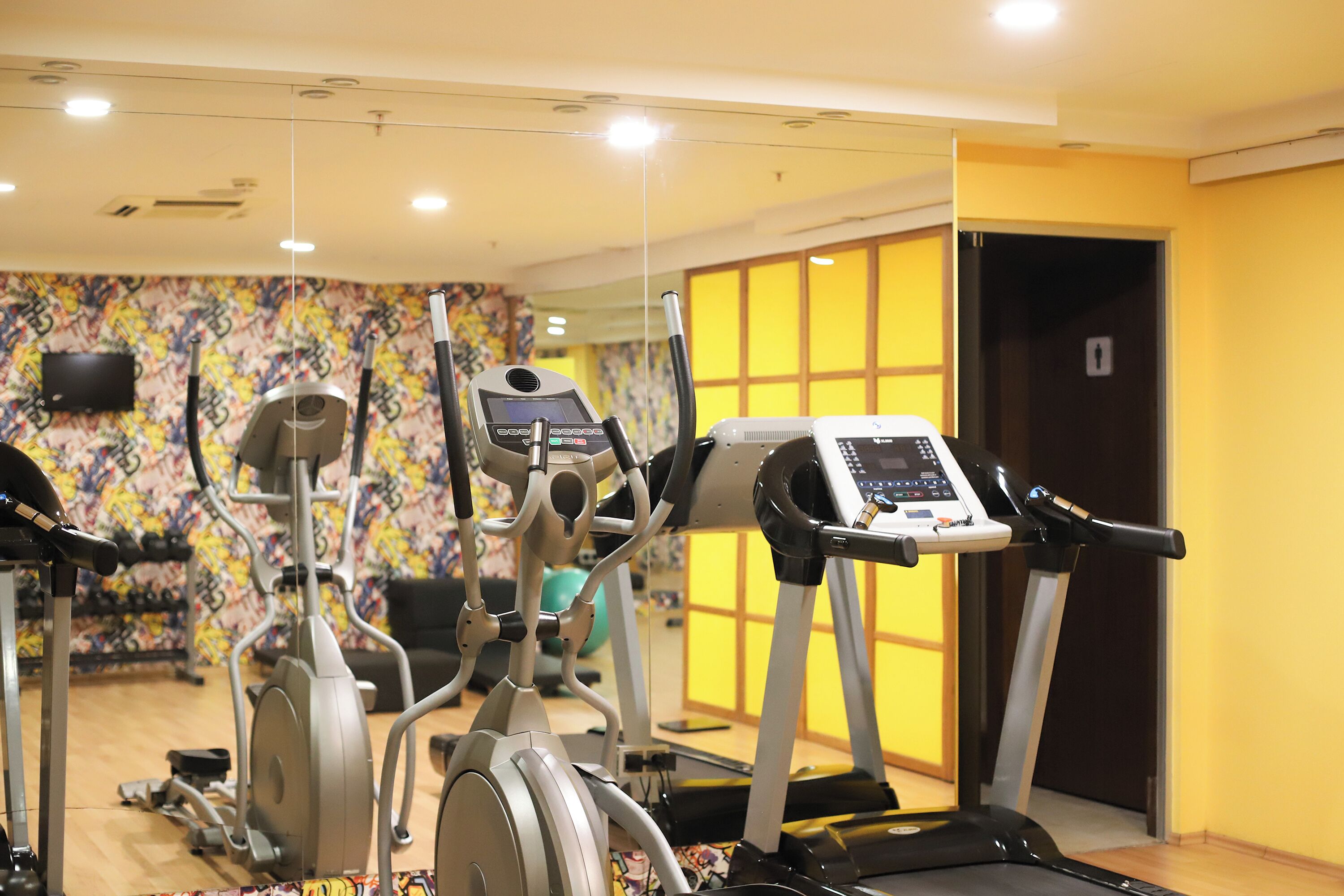 A gym with various exercise machines, a television mounted on the wall, and a yellow wall with a window.