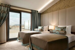 Ein Hotelzimmer mit zwei Betten, einem großen Fenster mit Vorhängen, Blick auf die Stadt und einem gemusterten Teppich.