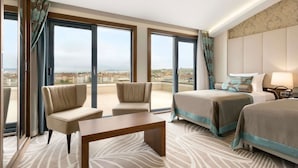 Habitación de hotel con dos camas, un sofá, una mesa y ventanas amplias con vista a la ciudad.