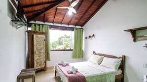 A bedroom with a bed, a wooden wardrobe, a small table, a ceiling fan, and a window with curtains. - Pousada NOVA ESTRELA INN (Búzios)