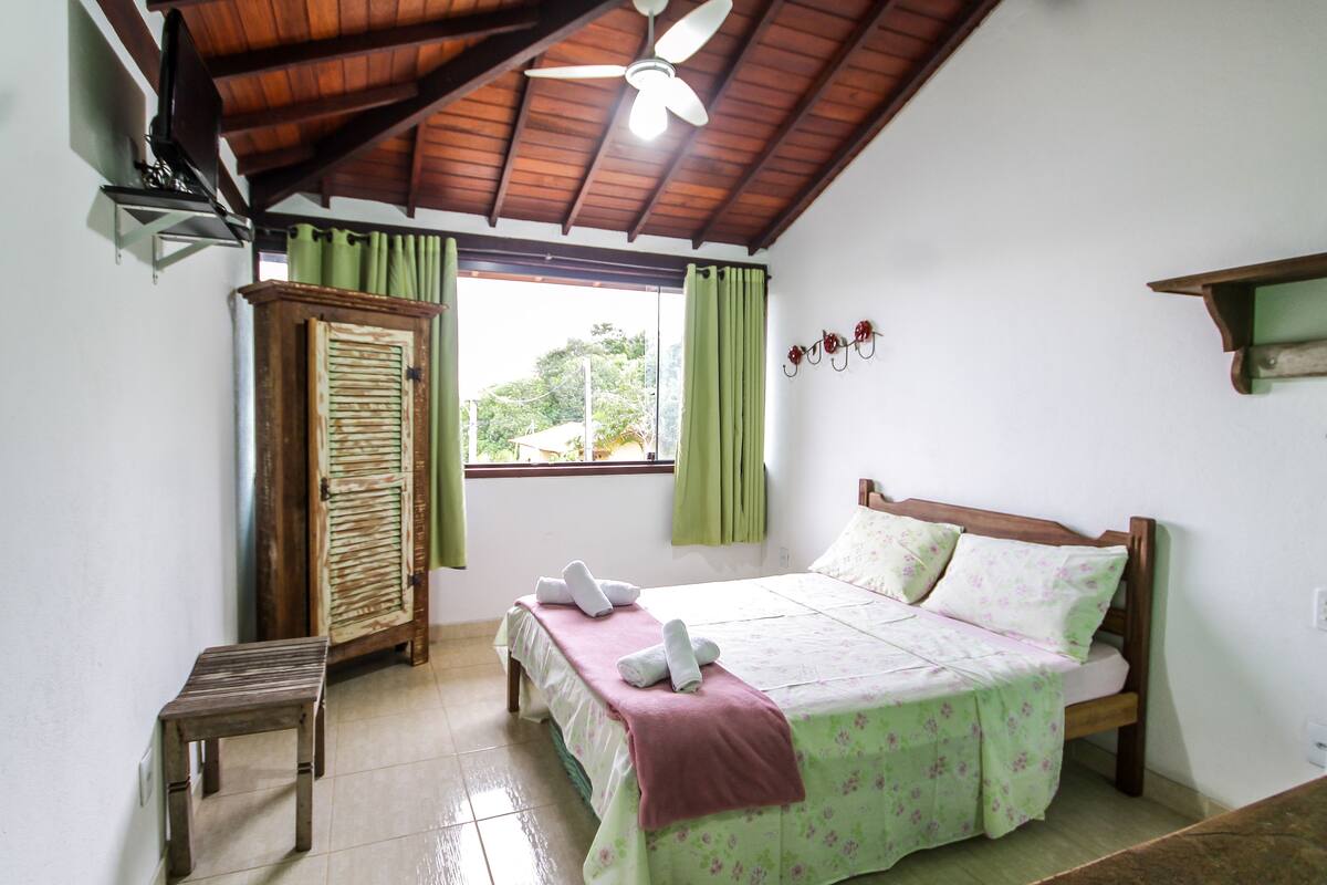 A bedroom with a bed, a wooden wardrobe, a small table, a ceiling fan, and a window with curtains.